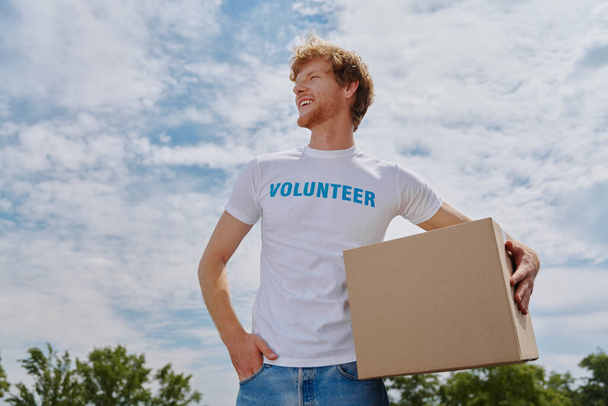 Confident young man in volunteer shirt carrying box and smiling while standing outdoors - 写真・画像