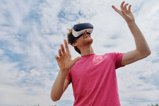 Handsome young redhead man in virtual reality headset gesturing against sky outdoors - Foto, imagen