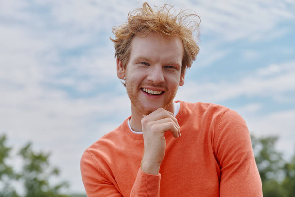Cheerful young redhead man holding hand on chin and smiling while standing outdoors - Фото, изображение