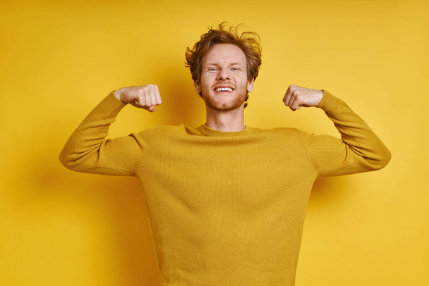 Cheerful redhead man showing his biceps while standing against yellow background - Valokuva, kuva