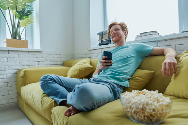 Cheerful young man watching TV and enjoying snacks while sitting on the couch at home - Foto, Imagen