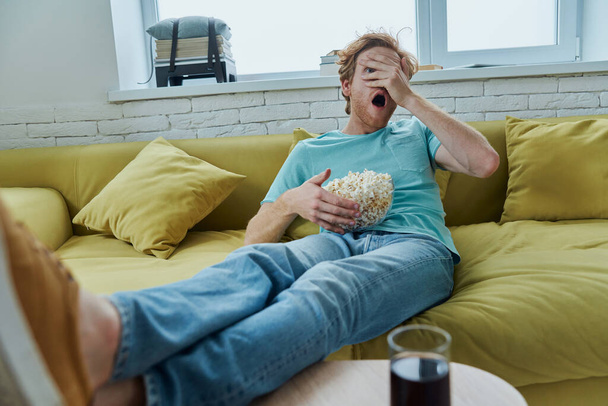 Shocked young man watching scary movie while sitting on the couch at home - Foto, Imagen