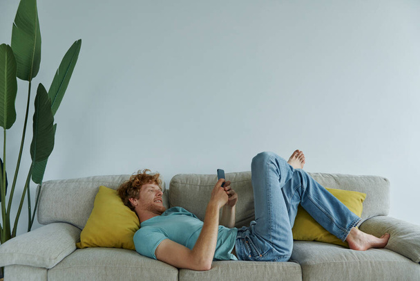Relaxed redhead man using smart phone while lying on the couch at home - Photo, image