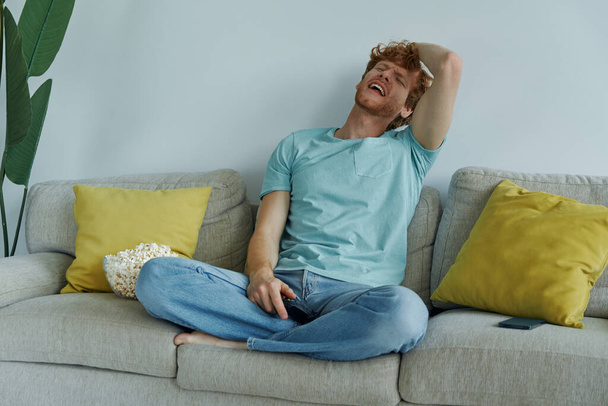 młody rudowłosy mężczyzna patrząc zdenerwowany podczas gry wideo na kanapie w domu - Zdjęcie, obraz
