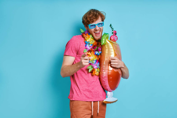 Happy man in Hawaiian necklace carrying cocktail shaped balloon against blue background - Foto, afbeelding