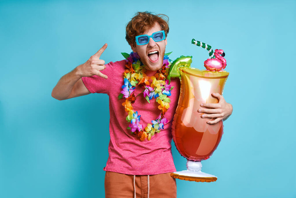Playful redhead man carrying cocktail shaped balloon and gesturing against blue background - Fotoğraf, Görsel