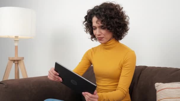 Latynoska kręcona kobieta grająca emocjonalnie w gry wideo na tablecie, siedząca na kanapie w domu. Pozytywne kobiety Ameryki Łacińskiej intensywnie grają w gry wyścigowe na tablecie w żółtym golfie - Materiał filmowy, wideo