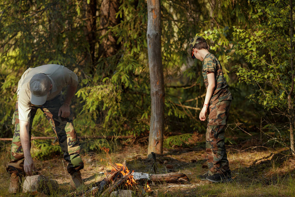 軍の服を着た息子と父は森の中でキャンプファイヤーをしています。冒険、旅行、観光、キャンプ、生存と避難の概念. - 写真・画像