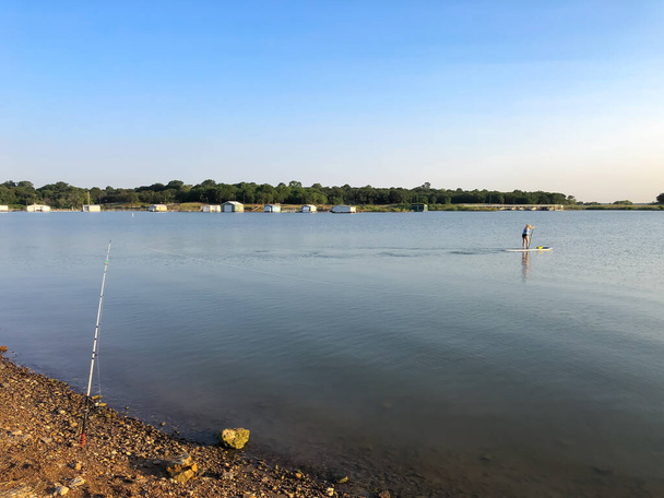 Temprano en la mañana en el lago con caña de pescar en portador de caña y mujer sana en el paddleboard de pie en Grapevine Lake, norte de Texas, Estados Unidos. Actividades recreativas de verano - Foto, imagen
