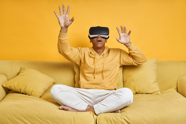 Excited young man in virtual reality glasses relaxing on the couch with yellow background - Fotó, kép