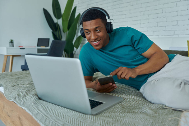 Cheerful African man in headphones using technologies while lying on bed at home - Photo, Image