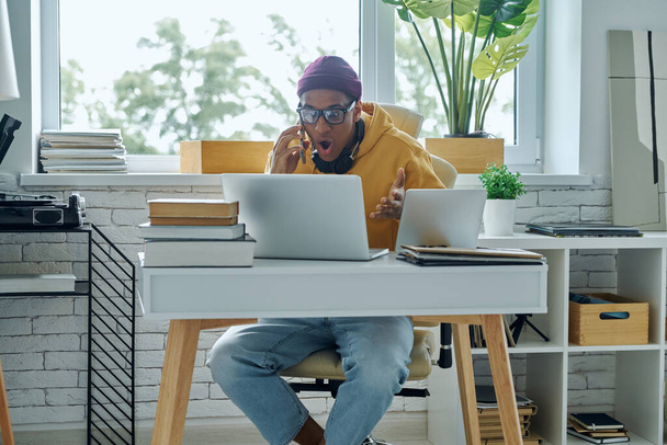 Surprised African man talking on mobile phone while working in home office - Fotó, kép