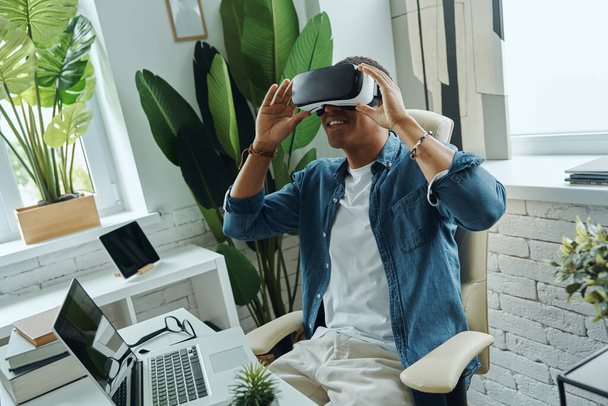 Cheerful African man wearing virtual reality glasses while sitting at the desk - Foto, Bild