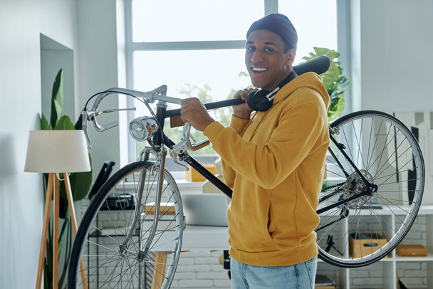 Happy young African man carrying bicycle on shoulder while standing in office - Foto, Bild