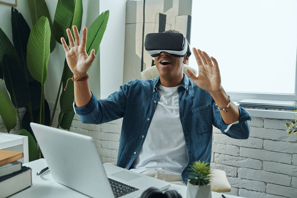 Happy African man wearing virtual reality glasses and gesturing while sitting in office - Fotó, kép