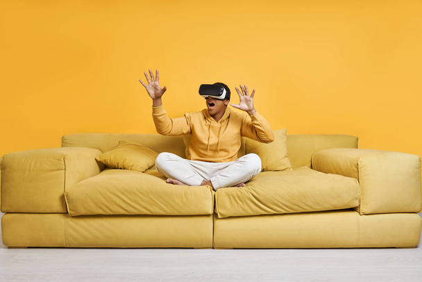 Excited man in virtual reality glasses gesturing while sitting on the couch with yellow background - Photo, Image