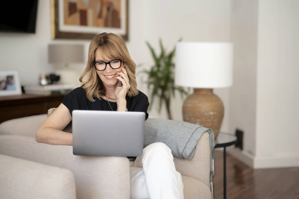 Confident woman using her laptop and mobile phone while sitting at the armchair and working from home. Home office. - Photo, Image
