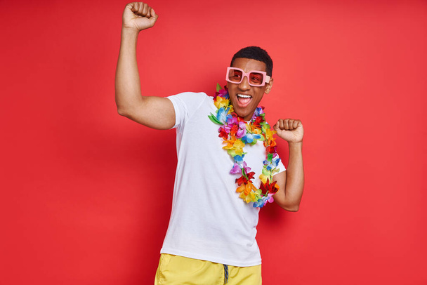 Handsome young man in Hawaiian necklace dancing against red background - Фото, изображение