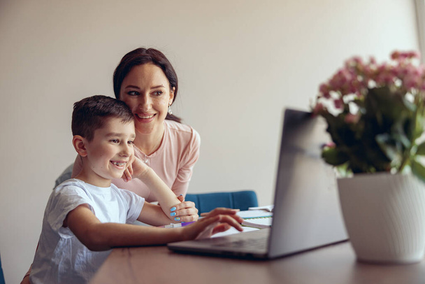 Kis aranyos iskolás fiú fogszabályzóval használja a laptopot az online tanulásban, boldog anya mosolyog. Szülőség. - Fotó, kép