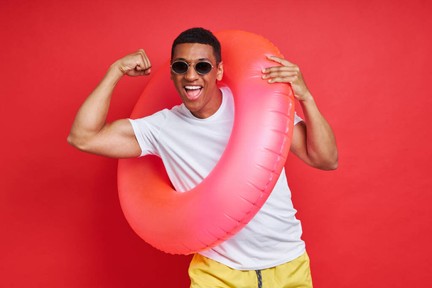 Playful African man carrying inflatable ring and showing bicep against red background - Фото, изображение