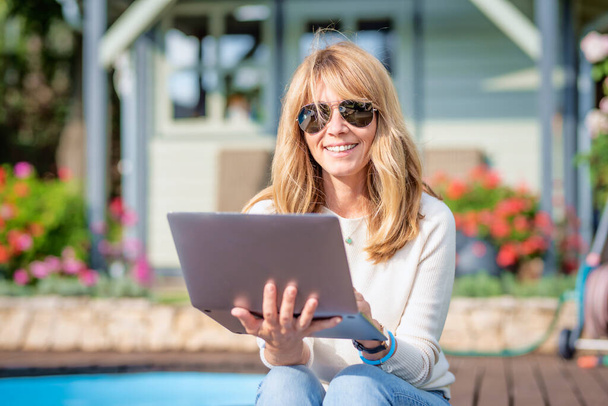 Attractive middle aged woman using laptop while sitting at the backyard at home and working. Home office.  - Foto, imagen