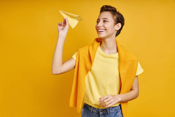 Happy young woman holding paper airplane while standing against yellow background - Foto, immagini