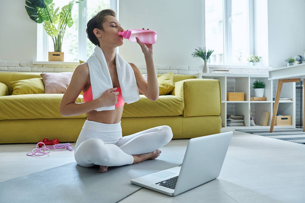 Confident young woman in sports clothing drinking water while sitting on exercise mat at home - Photo, image