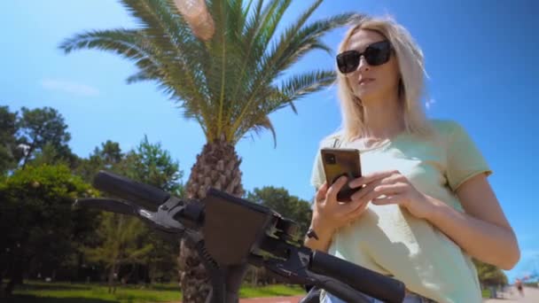 Woman puts smartphone to modern urban electric scooter, intending to unlock it. Modern urban transport. uses eco-transport in city center. palm trees, resort. party or weekend at sea. - 映像、動画