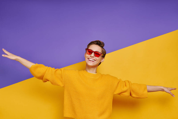 Playful young woman in eyeglasses keeping arms outstretched while standing against colorful background - Photo, image