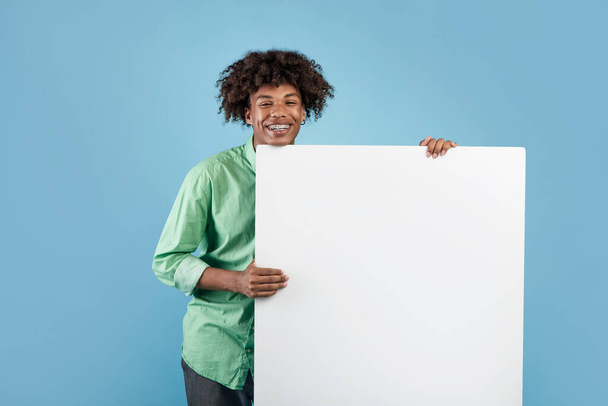 Mavi stüdyo arka planında reklam tasarımın için elinde boş bir poster tutan mutlu zenci genç adam. Heyecanlı Afrikalı Amerikalı genç boş afiş sunuyor. - Fotoğraf, Görsel