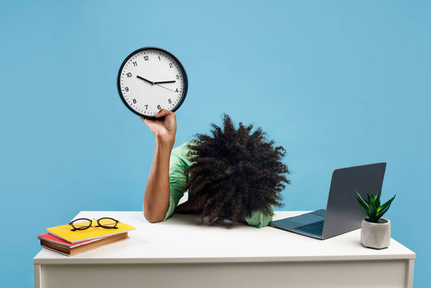 Müder schwarzer Student liegt mit Laptop und Büchern am Schreibtisch, hält Uhr und Kopf auf der Hand, fühlt sich vor blauem Studiohintergrund erschöpft - Foto, Bild