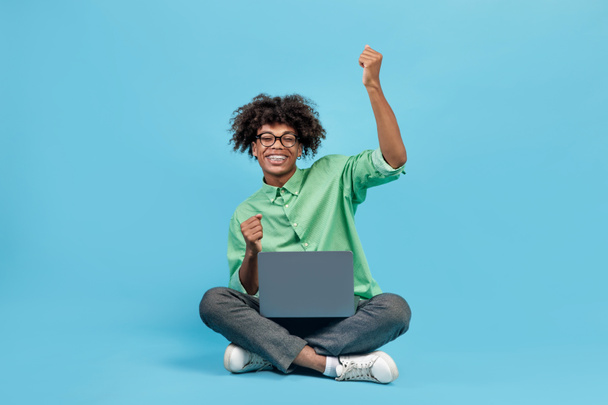Aufgeregter schwarzer Typ mit Laptop und zitternden Fäusten vor Freude, der auf blauem Studiohintergrund sitzt und in die Kamera lächelt. Tolle Online-News, bestandenes Prüfungskonzept - Foto, Bild