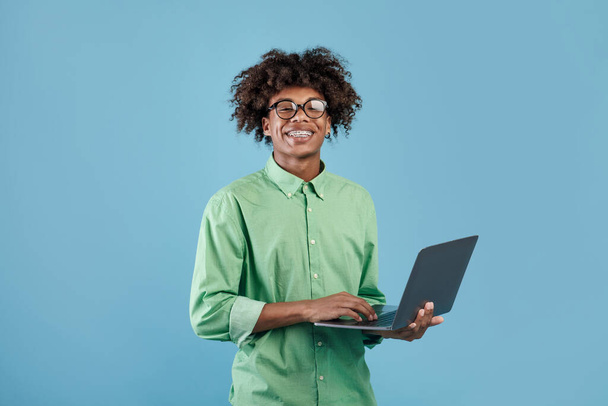 Θετικός μαύρος έφηβος χρησιμοποιεί το νέο του φορητό υπολογιστή, πληκτρολογεί στο πληκτρολόγιο και χαμογελάει στην κάμερα, ποζάρει πάνω από το μπλε φόντο του στούντιο. Gadgets, τεχνολογία και εκπαίδευση - Φωτογραφία, εικόνα