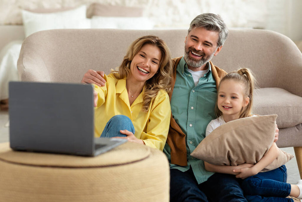 Ευτυχισμένη οικογένεια βλέποντας ταινία σε απευθείας σύνδεση στο φορητό υπολογιστή κάθεται στο σπίτι. Μεσήλικες γονείς και μικρή κόρη περνούν χρόνο μαζί το Σαββατοκύριακο. Επιλογή εστίασης - Φωτογραφία, εικόνα