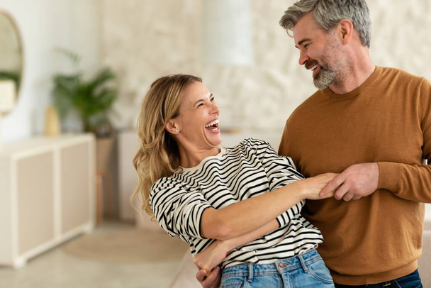 Szczęśliwego związku. Radosni małżonkowie w średnim wieku tańczą w nowoczesnym salonie w domu. Para bawiąca się i śmiejąca w domu. Małżeństwo i romans - Zdjęcie, obraz