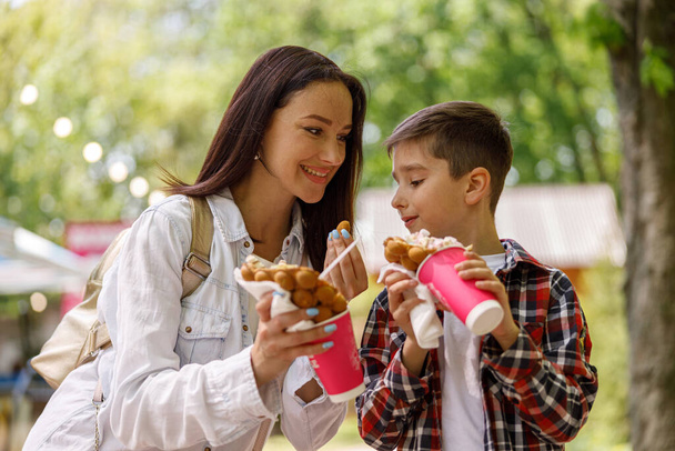 Küçük oğluyla parkta yürüyen ve tatlı waffle yiyen sevimli bir anne. Yaz zamanı. Yaz yürüyüşü. - Fotoğraf, Görsel