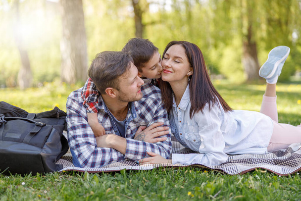 Parkta yeşil çimlere uzanmış çocuğu olan mutlu bir aile. Anne ve baba sarılıyor, oğlunu öpüyor. Piknik. - Fotoğraf, Görsel
