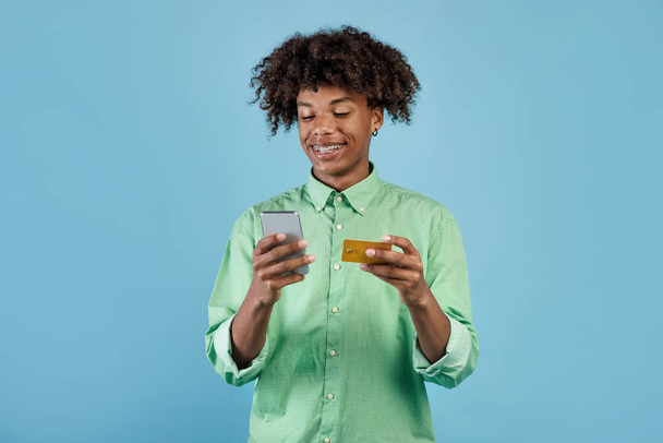 Szczęśliwy czarny facet trzyma komórkę i kartę kredytową, zakupy lub bankowość online, za pomocą nowej aplikacji mobilnej, pozowanie na tle niebieskiego studia. Koncepcja handlu elektronicznego - Zdjęcie, obraz