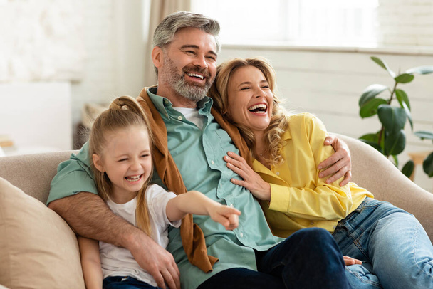 Padres de mediana edad abrazando a la pequeña hija riéndose ligándose pasando tiempo juntos sentados en un sofá en casa. Madre, padre y niño disfrutando el fin de semana. Concepto de estilo de vida familiar feliz - Foto, imagen