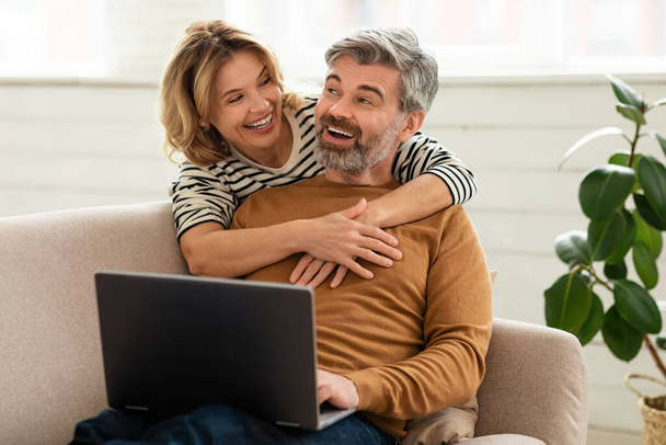 Cheerful Middle Aged Couple Using Laptop Together Browsing Internet And Watching Movie Online Sitting On Couch At Home. Wife Hugging Husband While Hes Working On Computer. Family And Technology - Photo, image