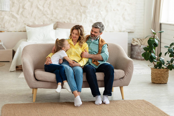 Семья из трех человек проводит время вместе, сидя и обнимая Бондинга в выходные позируя на диване дома. Родители обнимают своего ребенка. Happiness And Love, Parfelood - Фото, изображение