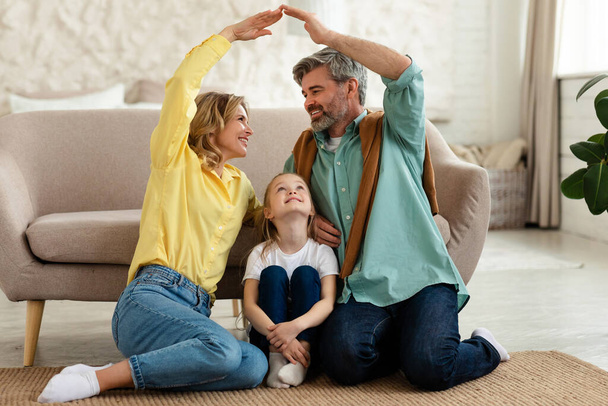 Family Housing. Cheerful Parents Joining Arms Making Symbolic Roof Above Their Little Daughter Sitting On Floor At Home. Dad, Mom And Child Dreaming About New House. Real Estate Concept - Photo, Image
