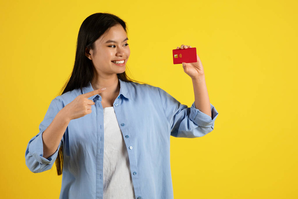Χαρούμενη, χαρούμενη, νεαρή Γιαπωνέζα που δείχνει με το δάχτυλο την πιστωτική της κάρτα, συστήνει εξοικονόμηση χρημάτων, απομονωμένη σε κίτρινο φόντο, στούντιο. Shopaholic και ψώνια, πώληση, χρηματοδότηση και πληρωμή αγορών - Φωτογραφία, εικόνα