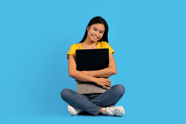 Vrolijke duizendjarige Aziatische vrouw in geel t-shirt zittend op de vloer, knuffelende laptop, geïsoleerd op blauwe achtergrond, studio. Moderne technologie, werk en studie op afstand, blog, goede advertentie, verkoop en aanbod - Foto, afbeelding