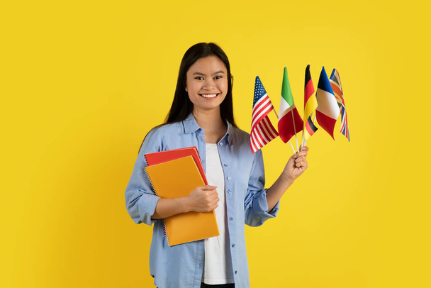 多くの国際旗を持つ若いアジアの女性学生や教師を満足させ、ノートブックは黄色の背景、スタジオに隔離されたカメラを見ています。現代の勉強と教育、学校で新しい言語を学ぶ - 写真・画像