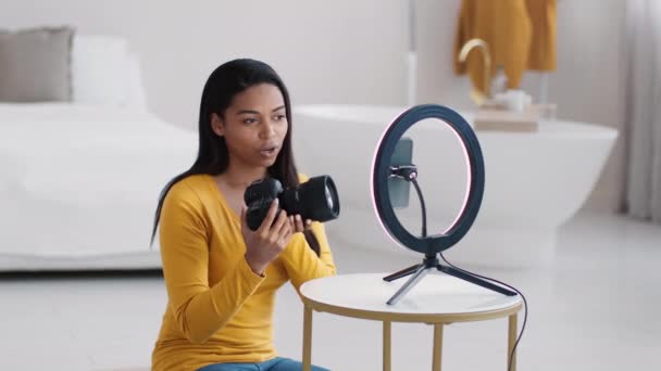 Professional black woman photographer recording video review about new photo camera, talking to subscribers via smartphone at home, slow motion - Footage, Video