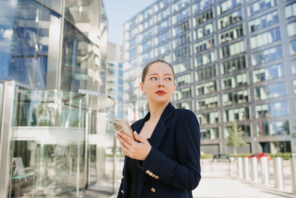 Una foto ravvicinata di un'agente immobiliare che sta usando un cellulare mentre aspetta. Una donna tra gli uffici del distretto finanziario. Una donna d'affari in un blazer nel gruppo di edifici alti. - Foto, immagini