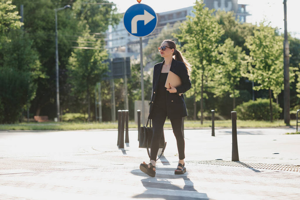 Una agente inmobiliaria femenina en una chaqueta azul marino sostiene una computadora portátil mientras cruza la calle en el centro de la ciudad. Una mujer de negocios está paseando cerca de la línea de árboles. - Foto, Imagen