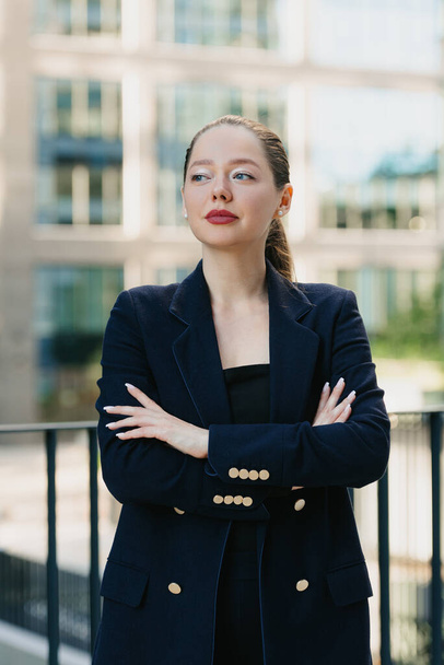 A female realtor in a blazer is standing with her arms crossed on her chest in a modern urban space in the financial district. A businesswoman in the contemporary garden in a cluster of tall buildings. - Fotó, kép