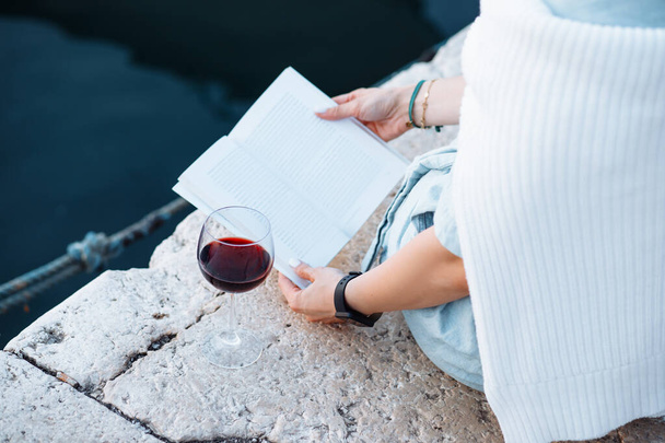 Frau in hellblauer Kleidung und gestricktem Baumwollpullover, der über den Schultern drapiert ist, sitzt mit Buch in der Hand auf einem Steg, neben ihr ein Glas Rotwein..  - Foto, Bild